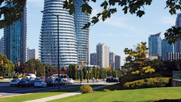 A city street in Mississauga lined with modern high-rise buildings, green space, and cars on a sunny day