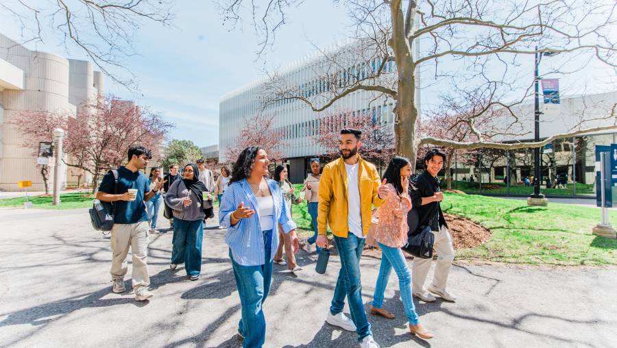 A group of prospective students walking together on a sunny day, chatting and smiling as they pass modern academic buildings and budding trees