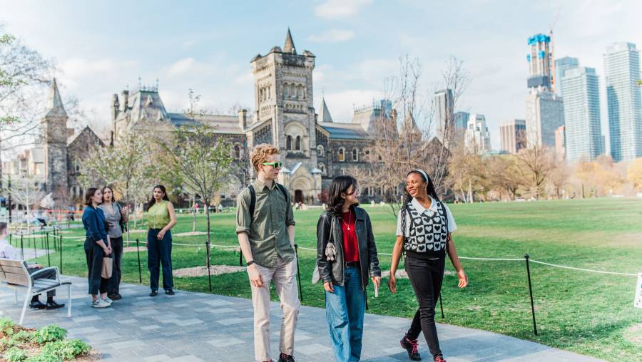 Three students walking on a path on a sunny day with more students and a historic looking building in the background