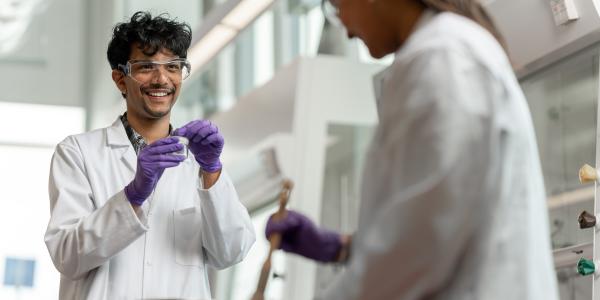 Two students in a lab wearing white lab coats; one is smile, wearing gloves and holding a vial
