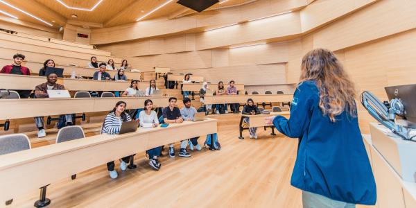 A person speaks to a group inside a modern lecture hall, with students seated and listening from tiered rows