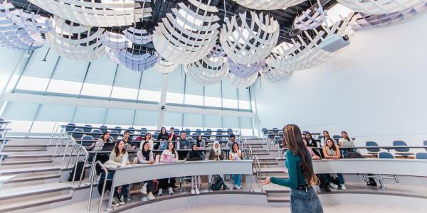 Professor speaks to a group in a modern lecture hall with sculptural ceiling panels.
