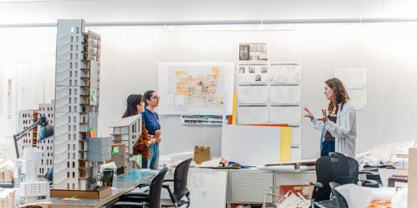 Students in an architecture studio discuss a project, surrounded by design models, drawings and planning materials