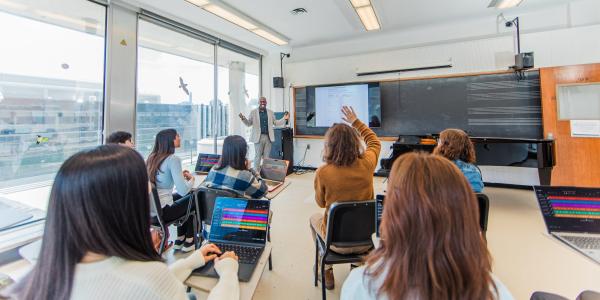 Professor lecturing in a small classroom with several students; one student is raising his hand to ask a question