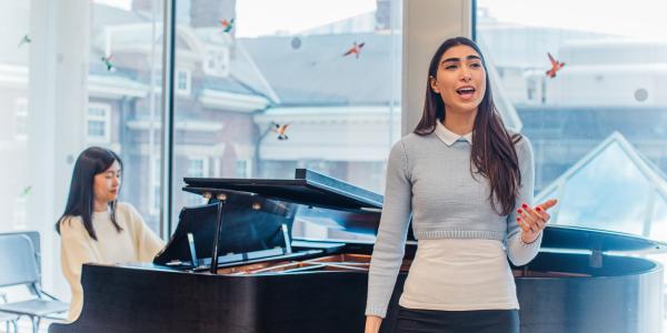 Student singing in a classroom with another student playing a grand piano behind her