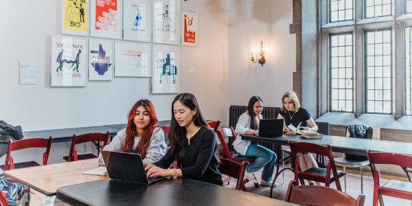 Students work on laptops in a high-ceilinged room with large windows and graphic artwork on the walls behind them