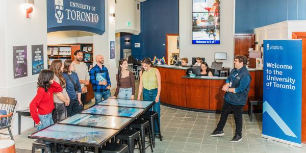 A tour guide welcomes a group of prospective students inside a visitor centre, with a blue banner that says “Welcome to the University of Toronto”