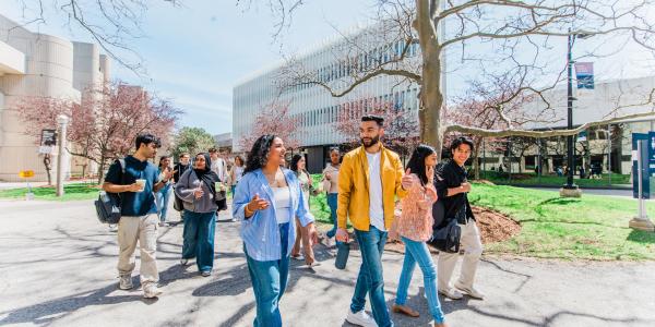 A group of prospective students walking together on a sunny day, chatting and smiling as they pass modern academic buildings and budding trees