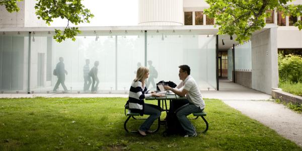 Two students sit at a table on a grassy lawn in front of a glass-walled campus building