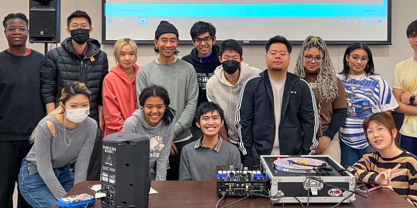 A diverse group of students posing in a classroom, gathered around DJ equipment and speakers