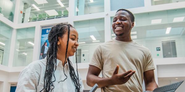 Two students smile and talk while holding a tablet in a bright, modern campus building