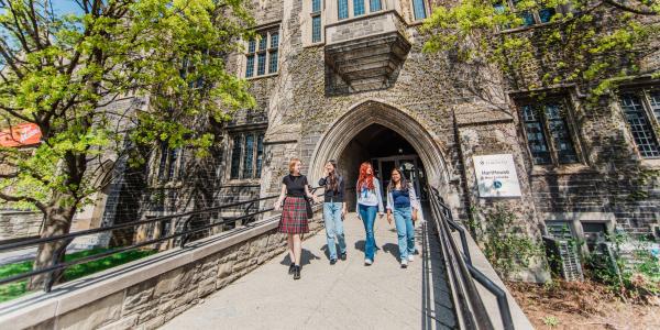 Four students walk into a historic stone building, under a stone archway framed by leafy trees on a sunny day