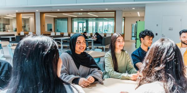 A group of students sitting around a table, talking and smiling in a bright, modern campus space