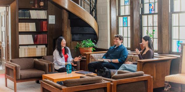 Three students sitting and talking in a wood-paneled study space with large stained-glass windows and a spiral staircase