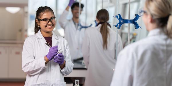 Student in a lab coat holding lab equipment, surrounded by classmates also wearing lab coats