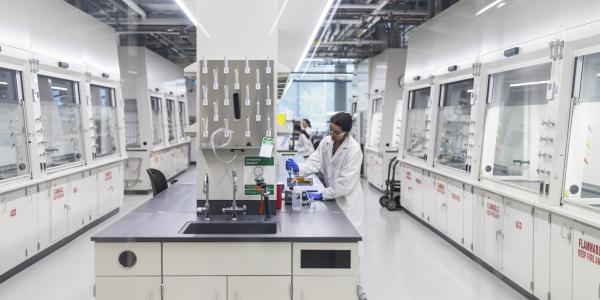 A student in a lab coat works at a station in a modern laboratory with fume hoods and scientific equipment