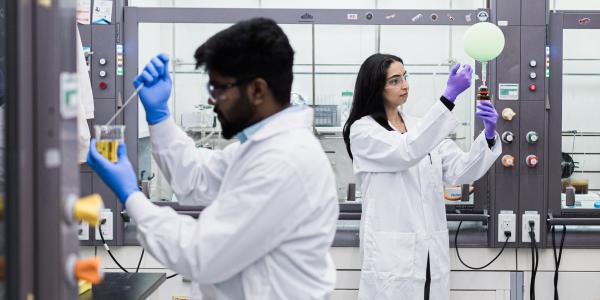 Two students in lab coats working in a chemistry lab, one seated using a pipette and the other standing near a fume hood