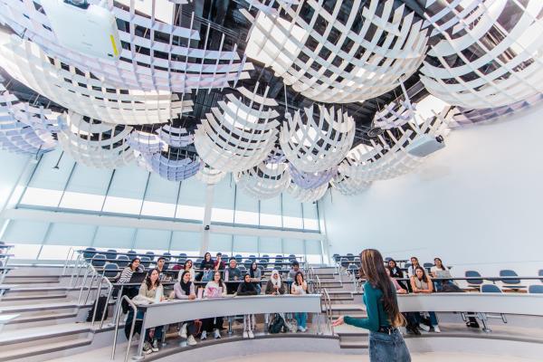 Professor speaks to a group in a modern lecture hall with sculptural ceiling panels.