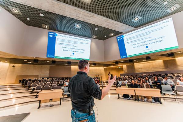 An instructor speaks to students in a lecture hall with large projection screens