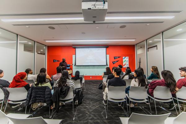 Students sitting in on an admissions presentation in a modern room, with a speaker presenting at the front and a screen