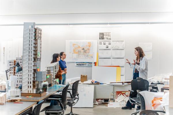 Students in an architecture studio discuss a project, surrounded by design models, drawings and planning materials
