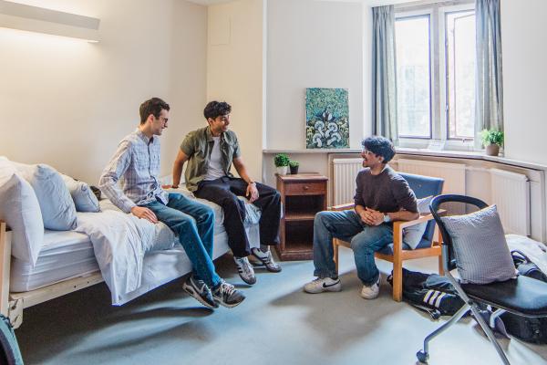 Two students sitting on a bed, another one of a chair, inside a nicely furnished residence room