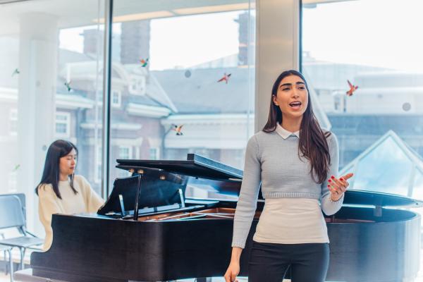 Student singing in a classroom with another student playing a grand piano behind her