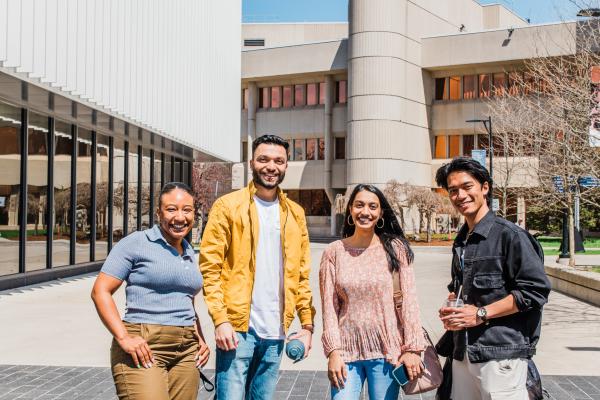 Four students smile for a photo outside academic buildings at University of Toronto Mississauga on a clear day
