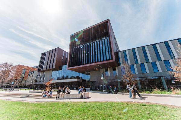 Students walk and sit outside a modern glass, metal and wood academic building on a sunny day