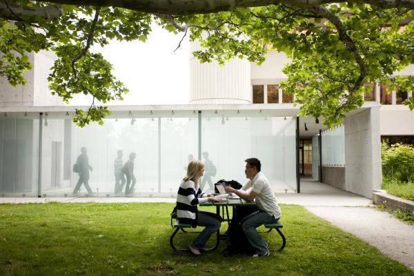 Two students sit at a table on a grassy lawn in front of a glass-walled campus building