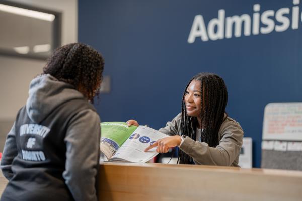 A staff member at in the admissions office smiles while assisting a student and pointing to information in a booklet