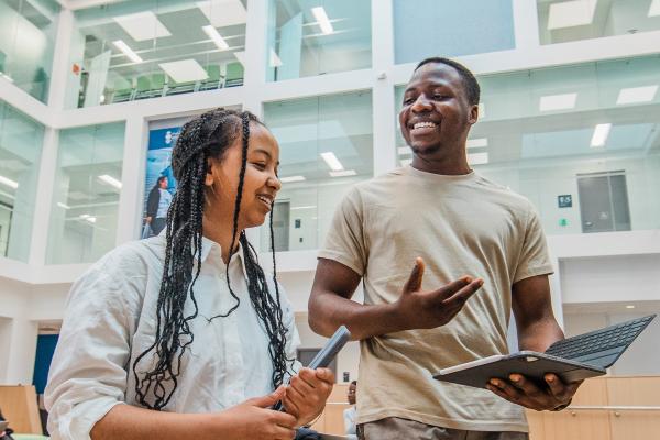 Two students smile and talk while holding a tablet in a bright, modern campus building
