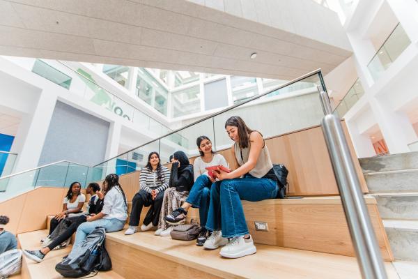 Students sitting and talking on tiered wooden seating inside a bright, modern building