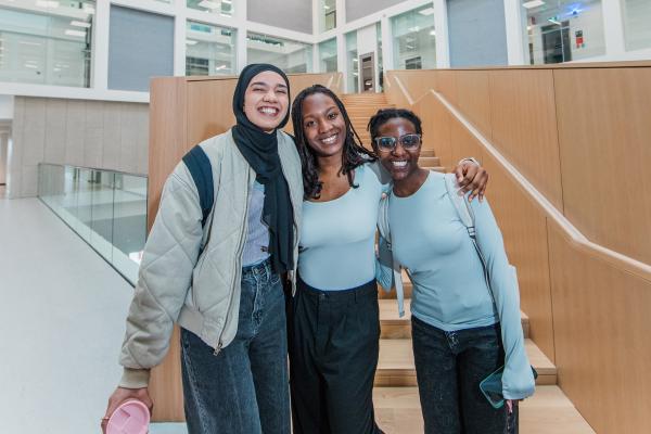Three smiling students standing together inside a bright, modern university building