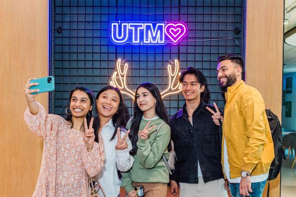 Group of five students taking a selfie in front of a neon UTM sign with a heart
