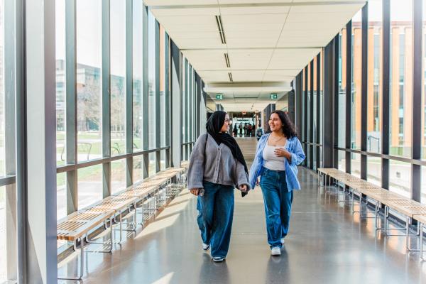 Two smiling students walk together through a glass-walled corridor