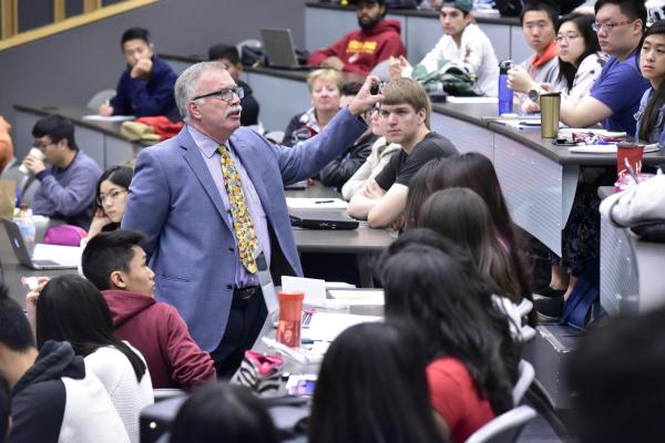 A professor wearing a blue blazer and yellow tie engages with students during a large lecture