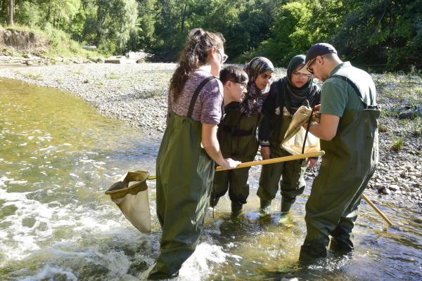 A group of students and a professor wearing waders stand in a shallow stream, examining samples