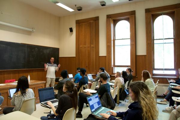 A professor in front of a chalkboard speaks to a class of students using laptops in a traditional, wood-paneled lecture hall