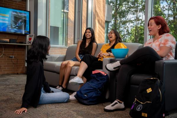 Group of four students sitting in a common room talking and laughing, with a TV on in the background