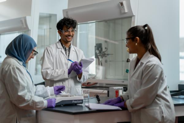 Three scientists in lab coats and safety gear working around a lab bench, with one taking notes