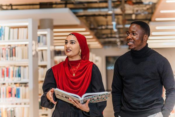 Two students walking together in a library. One of the students is holding an open book