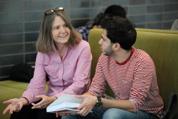 A student and a faculty member sitting on a green couch, having a conversation and holding a notebook