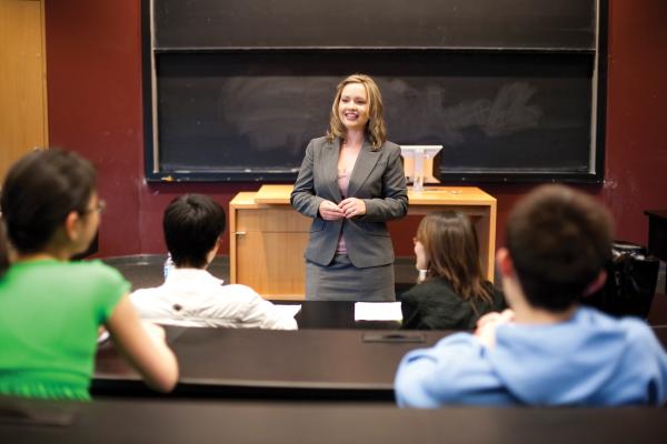 Professor wearing a grey blazer teaching a class, with a blackboard behind her