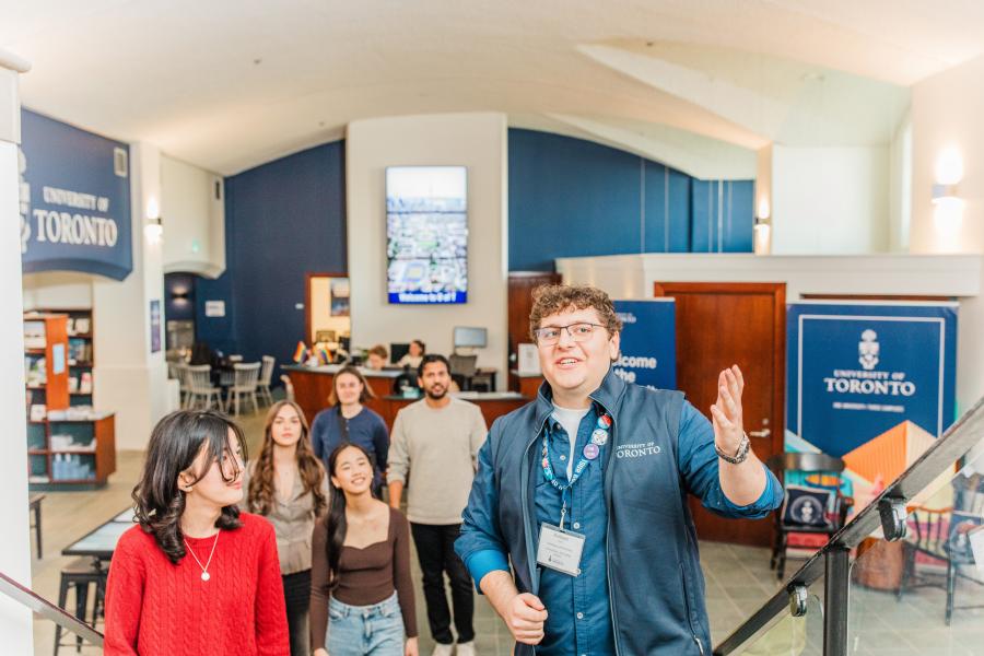 A student tour guide leads a group, with campus signage and visitors in the background