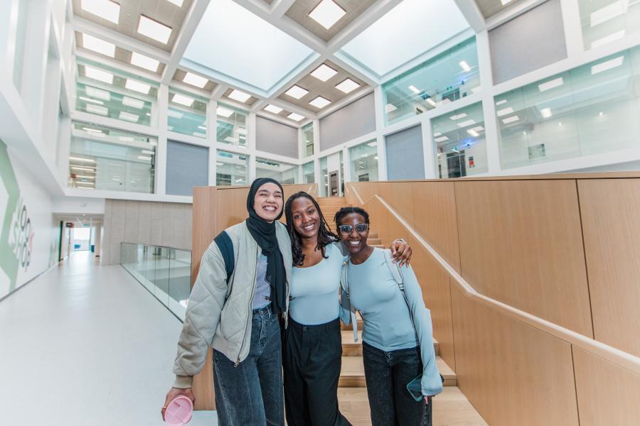 Three smiling students standing together inside a bright, modern university building