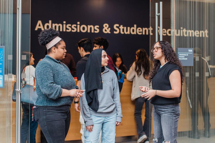 Three students talking in front of the Admissions & Student Recruitment office, with more people inside the office area