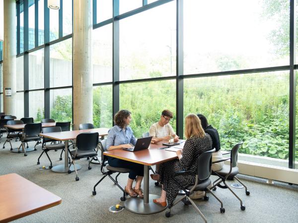 Students sitting at a table inside, next to a window-lined wall looking out lush greenery