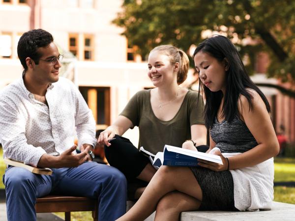 Three students sitting outside on campus, talking and studying with books