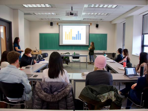 A student presents a bar chart on a screen to a class seated around tables